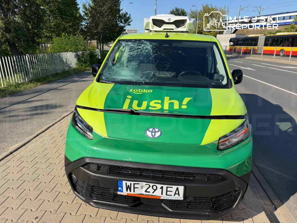 Zielony samochód dostawczy z logo "żabka" i napisem "iush!" z uszkodzoną szybą stoi na chodniku obok drogi.