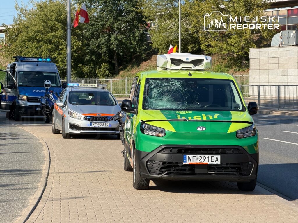 Zielony samochód dostawczy z uszkodzoną szybą stoi na chodniku, obok policyjne pojazdy z włączonymi sygnałami świetlnymi.
