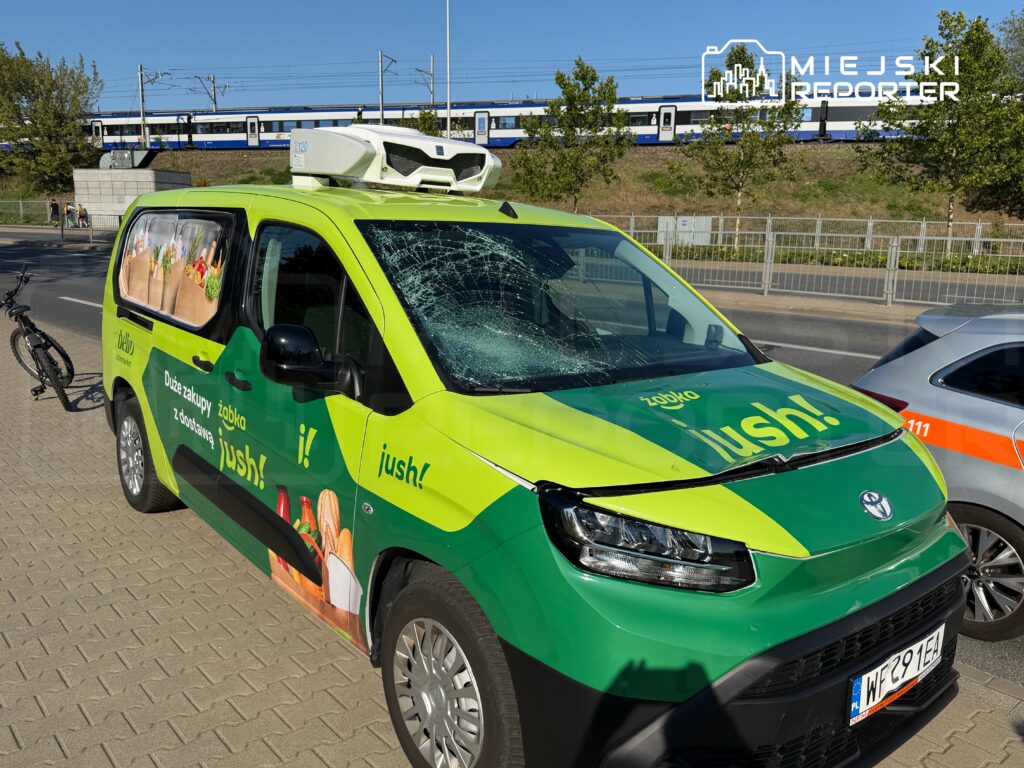 Zielony samochód dostawczy z reklamą usługi „jush!” stoi na chodniku, obok roweru, z pękniętą szybą czołową. W tle widoczny pociąg.