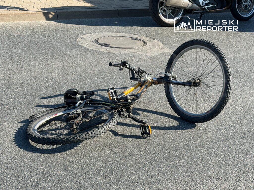 Rower leży na asfalcie, z jedną odkręconą kierownicą i uszkodzoną przednią oponą. W tle widoczna część motocykla.
