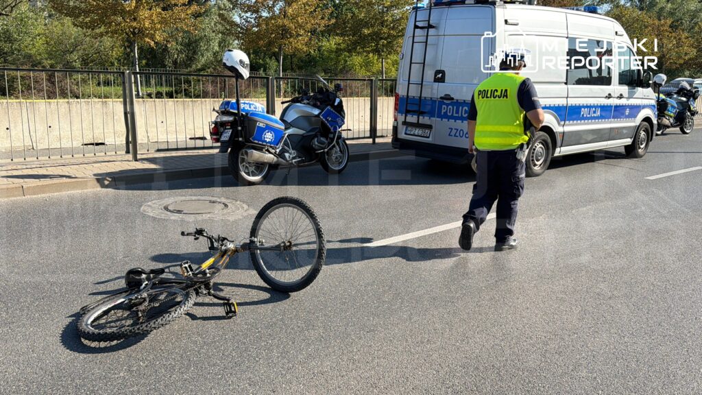 Na jezdni leży przewrócony rower, obok policjant w kamizelce odblaskowej rozmawia z innym funkcjonariuszem.