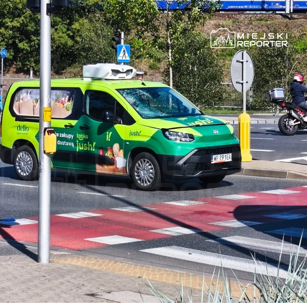 Zielony samochód dostawczy z reklamą usługi zakupów online przejeżdża przez skrzyżowanie z pasami dla pieszych.