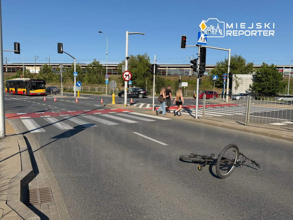 Rower leży na jezdni obok przejścia dla pieszych, a piesi przechodzą przez ulicę w pobliżu przystanku autobusowego.