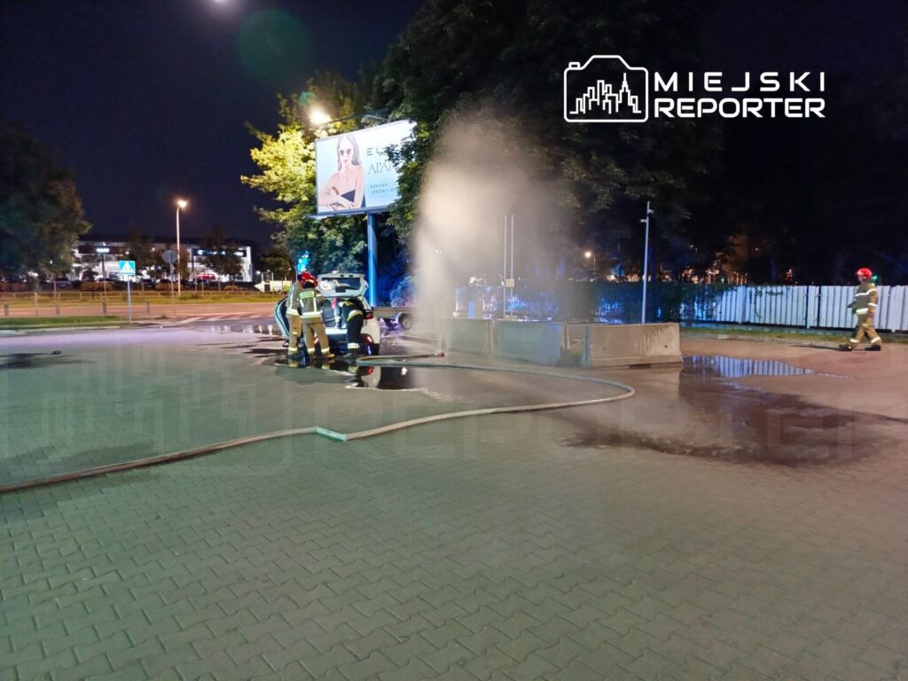 Grupa strażaków w mundurach gasi pożar w nocy, obok wody wypływającej z hydrantu na placu.