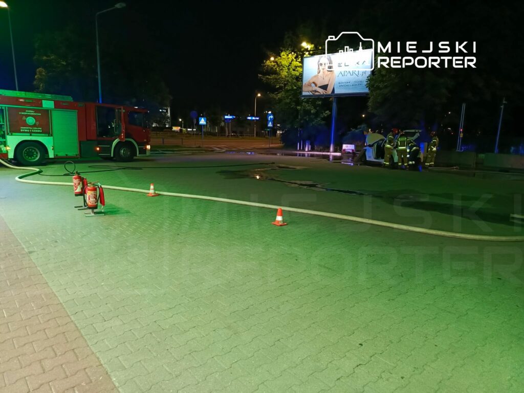 Strażacy pracują przy usunięciu skutków zdarzenia na drodze, obok wozu strażackiego i hydrantu.