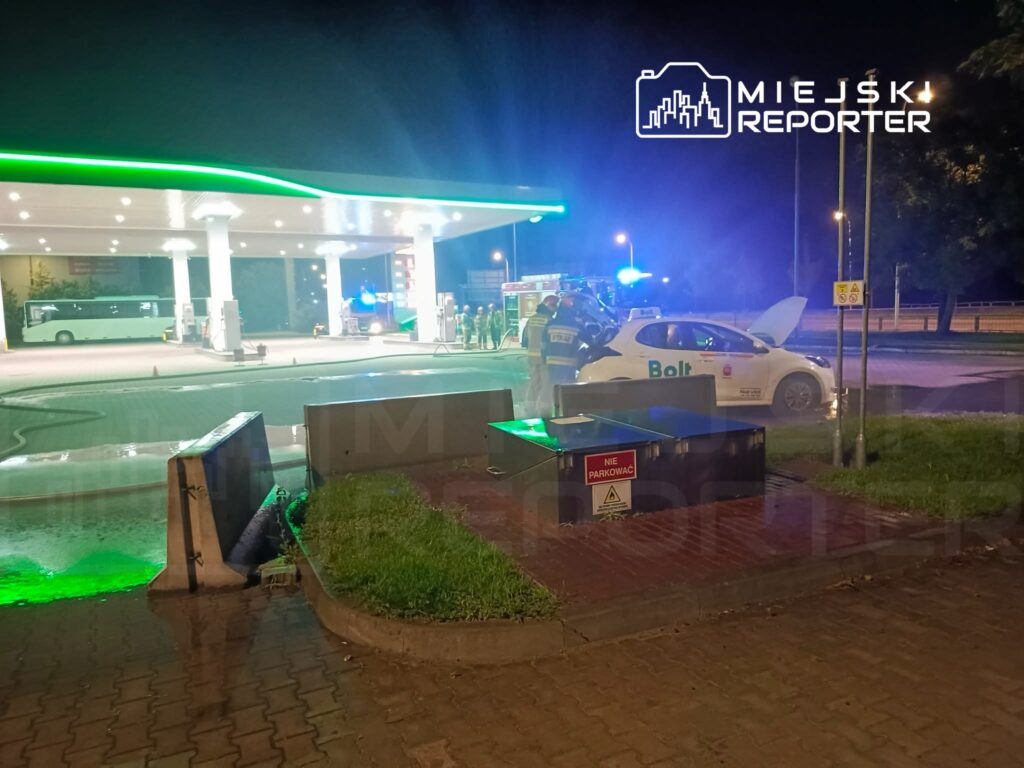 Grupa strażaków i policjantów pomaga kierowcy samochodu Bolt na stacji benzynowej w nocy. W tle widoczny autobus.
