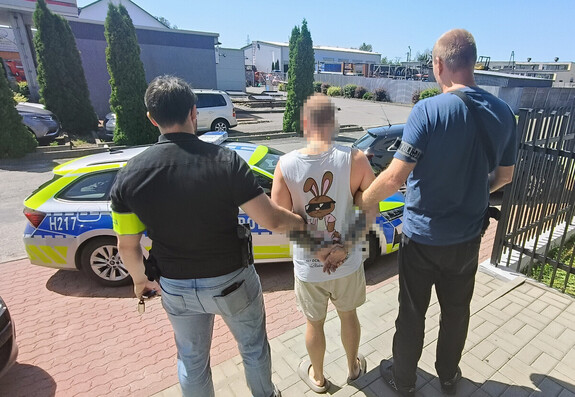Mężczyzna w krótkich spodniach, z rękami związanymi z tyłu, prowadzony przez dwóch funkcjonariuszy policji obok radiowozu.