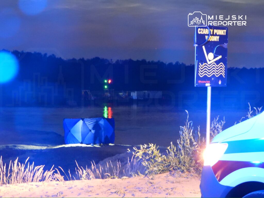 Na brzegu wody stoi niebieski namiot, a obok znajduje się znak informacyjny o punkcie wodnym. W tle widać reflektory i wodę odbijającą światło.