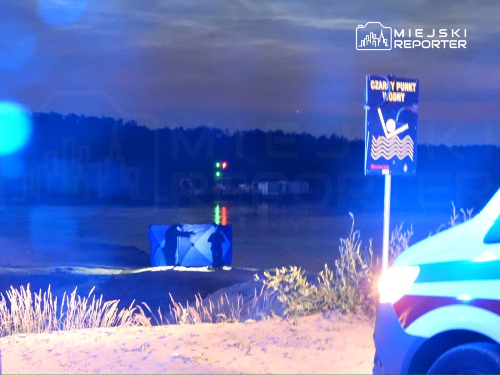 Mężczyźni w niebieskich ubraniach stoją przy namiocie ratunkowym na plaży, w tle widoczne światła policyjnych pojazdów.