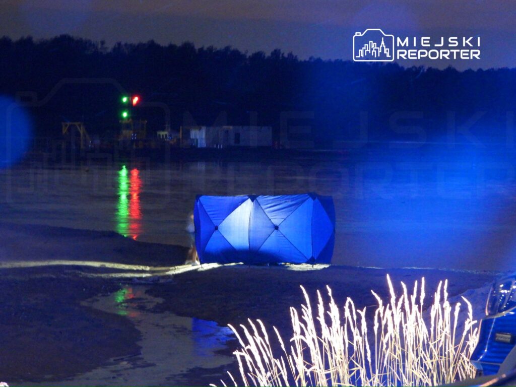 Na plaży stoi niebieski namiot, a w tle widać zielone i czerwone światła odbijające się w wodzie.