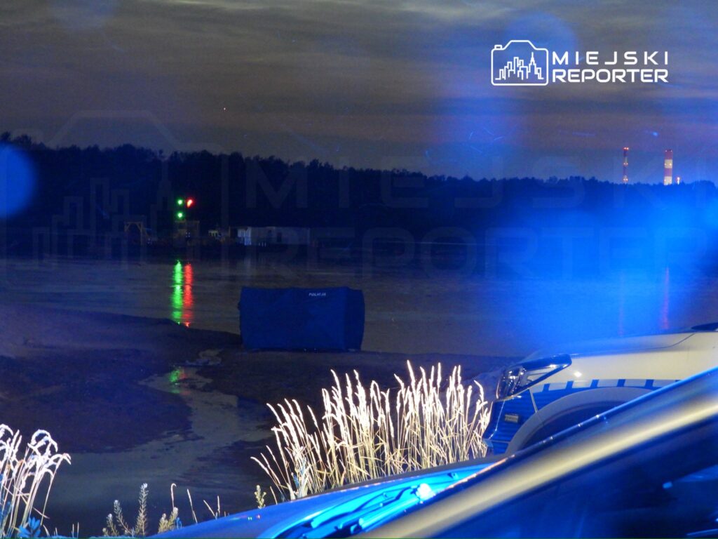 Policyjny namiot w ciemności, z niebieskimi światłami radiowozu, w tle woda i leśne otoczenie.