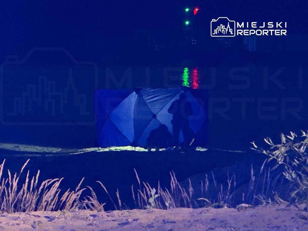 Mężczyzna przy namiocie na plaży, w tle odbicie świateł na wodzie, nocna sceneria.
