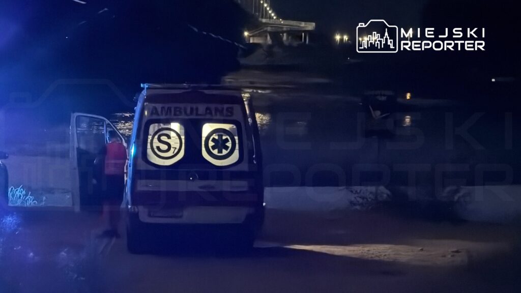 Ambulans z otwartymi drzwiami stoi nad brzegiem wody, w tle widać most i nocne oświetlenie.