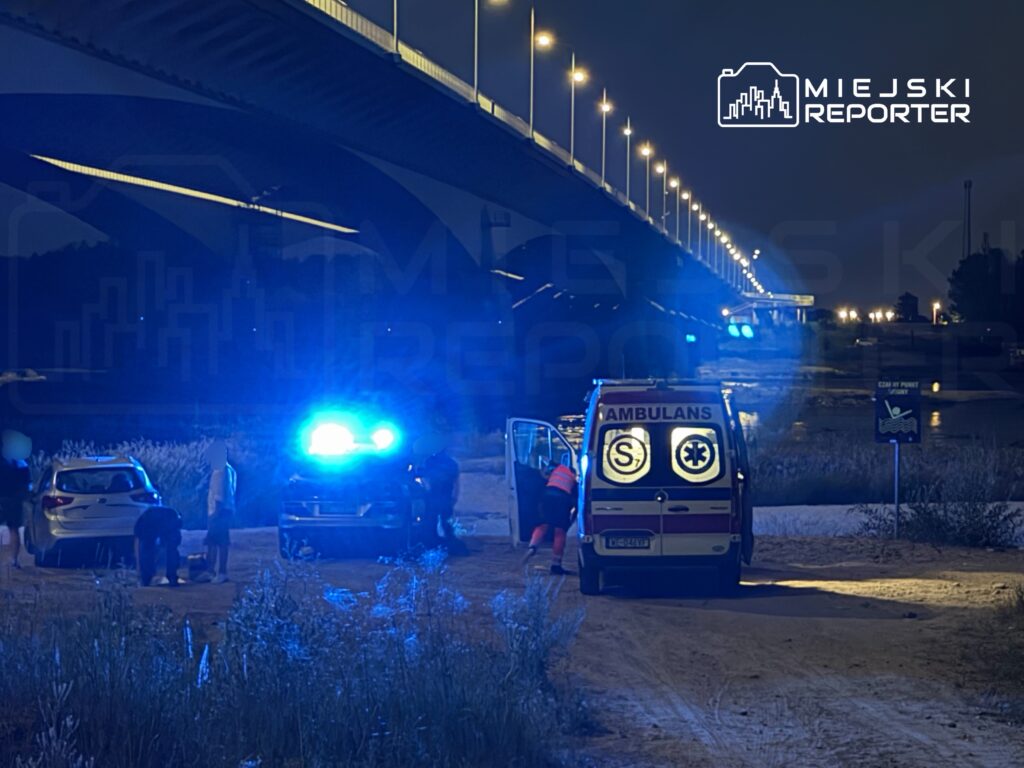 Grupa osób w ciemności otacza ambulans z włączonymi światłami, stojąc obok zaparkowanego samochodu.