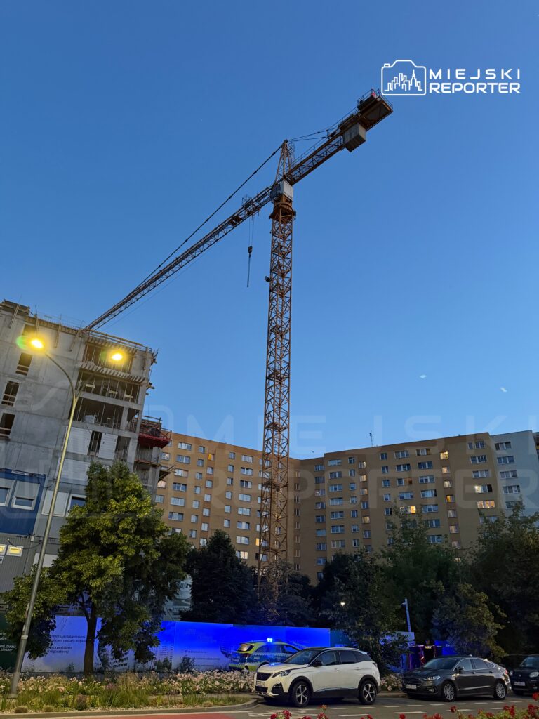Dźwig budowlany unoszący elementy konstrukcyjne obok nowego budynku mieszkalnego w wieczornym świetle.