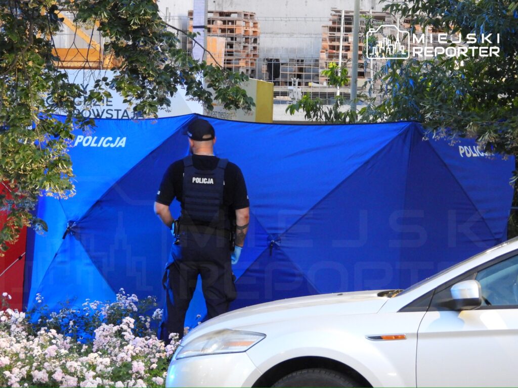 Policjant w kamizelce ochronnej stoi przy niebieskim namiocie, otoczony przez kwiaty, w tle budowa.