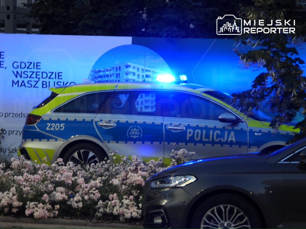Policyjny radiowóz z włączonymi sygnałami świetlnymi zaparkowany obok kwietników z różowymi kwiatami.