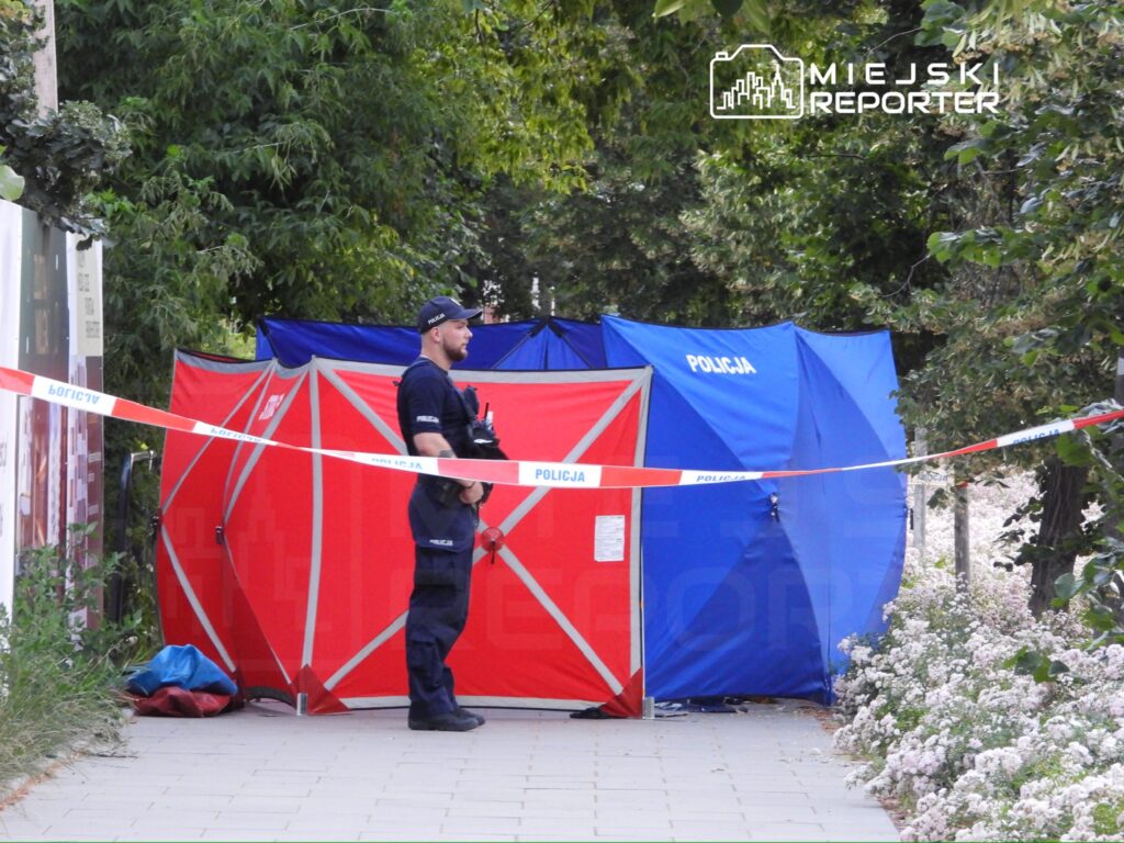 Policjant w mundurze stoi przy namiotach zabezpieczających miejsce zdarzenia, otoczony taśmą policyjną.