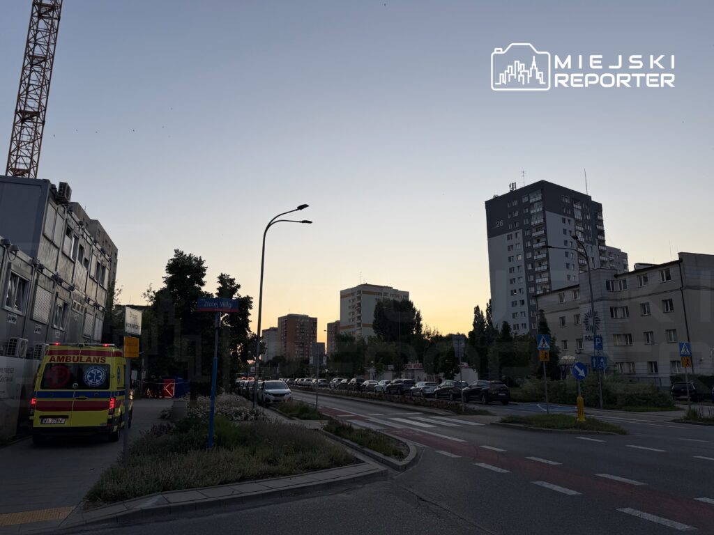 Ambulans zaparkowany przy ulicy, w tle widać nowoczesne budynki oraz zmieniające się kolory nieba o zachodzie słońca.