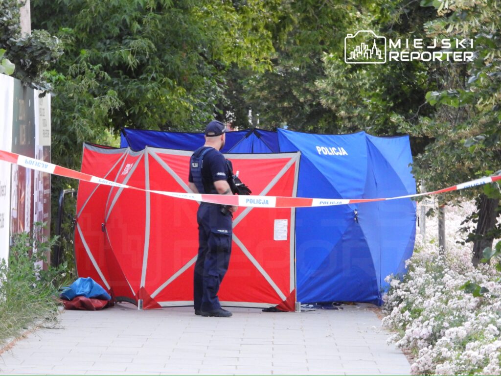 Policjant w mundurze obserwuje miejsce zdarzenia, gdzie rozstawione są namioty policyjne w otoczeniu zieleni.