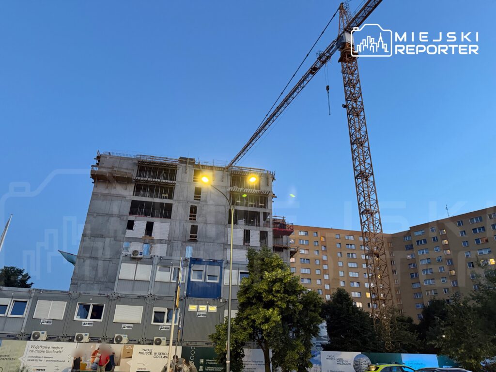 Nowy budynek w trakcie budowy z widocznym dźwigiem i oświetleniem ulicznym, w tle bloki mieszkalne.
