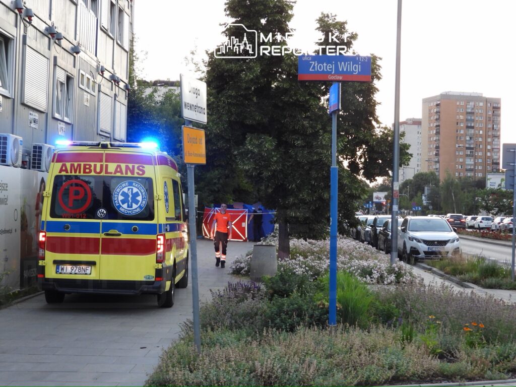 Ambulans z włączonymi sygnałami świetlnymi stoi przy drodze, obok pracownika w pomarańczowym ubraniu.