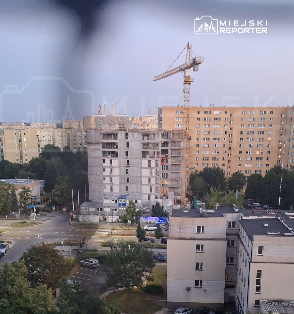 W tle widać budynek w trakcie budowy, z dźwigiem oraz otaczające go blokowiska mieszkalne. Na pierwszym planie znajduje się zieleń oraz miejsca parkingowe.