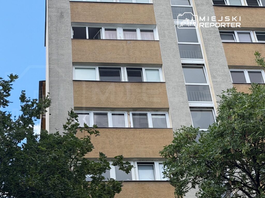 Wysoki blok mieszkalny z oknami w różnych kolorach, otoczony zielonymi drzewami i błękitnym niebem.