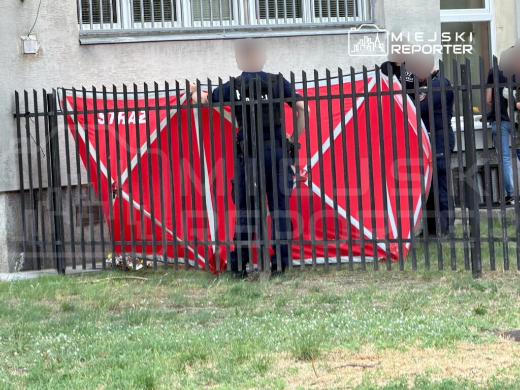 Strażacy ustawiają czerwoną osłonę w pobliżu ogrodzenia, w tle widoczne okna budynku.