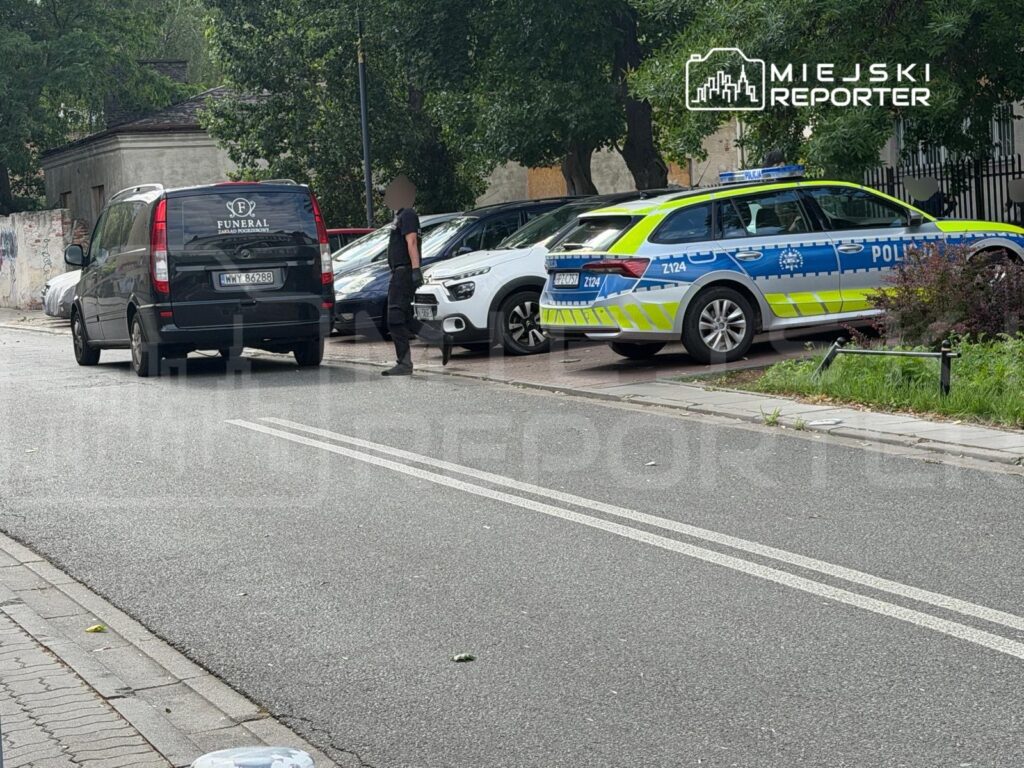 Mężczyzna w czarnym stroju spaceruje obok furgonetki z napisem „FUNERAL” oraz policyjnego auta na ulicy.