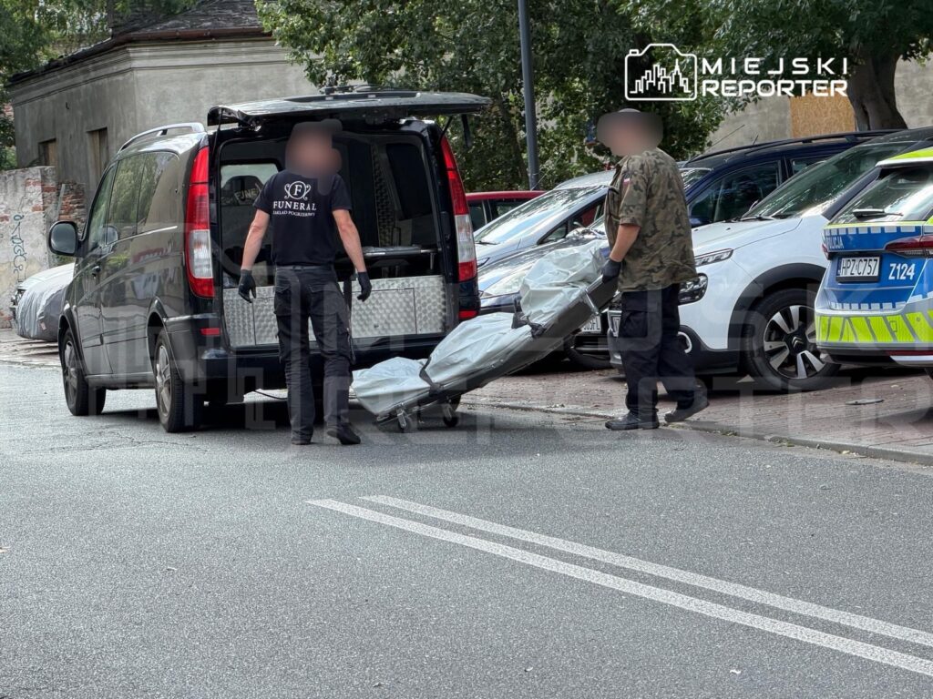 Dwaj pracownicy w czarnych ubraniach transportują przykryty obiekt z furgonetki na ulicy.