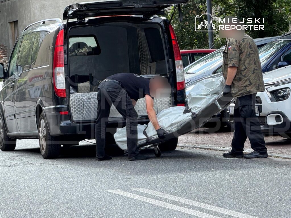 Dwaj mężczyźni w rękawicach wynoszą zabezpieczony przedmiot z czarnego vana na ulicy.