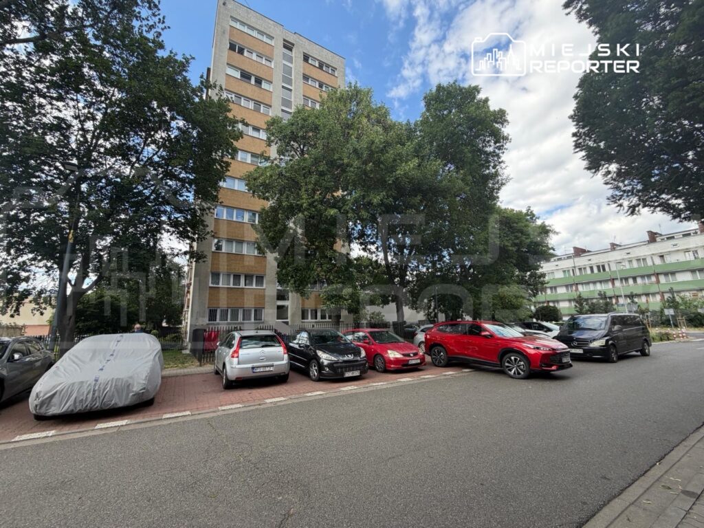 Wysoki budynek mieszkalny otoczony drzewami, zaparkowane samochody na ulicy, w tym przykryty pokrowcem pojazd.