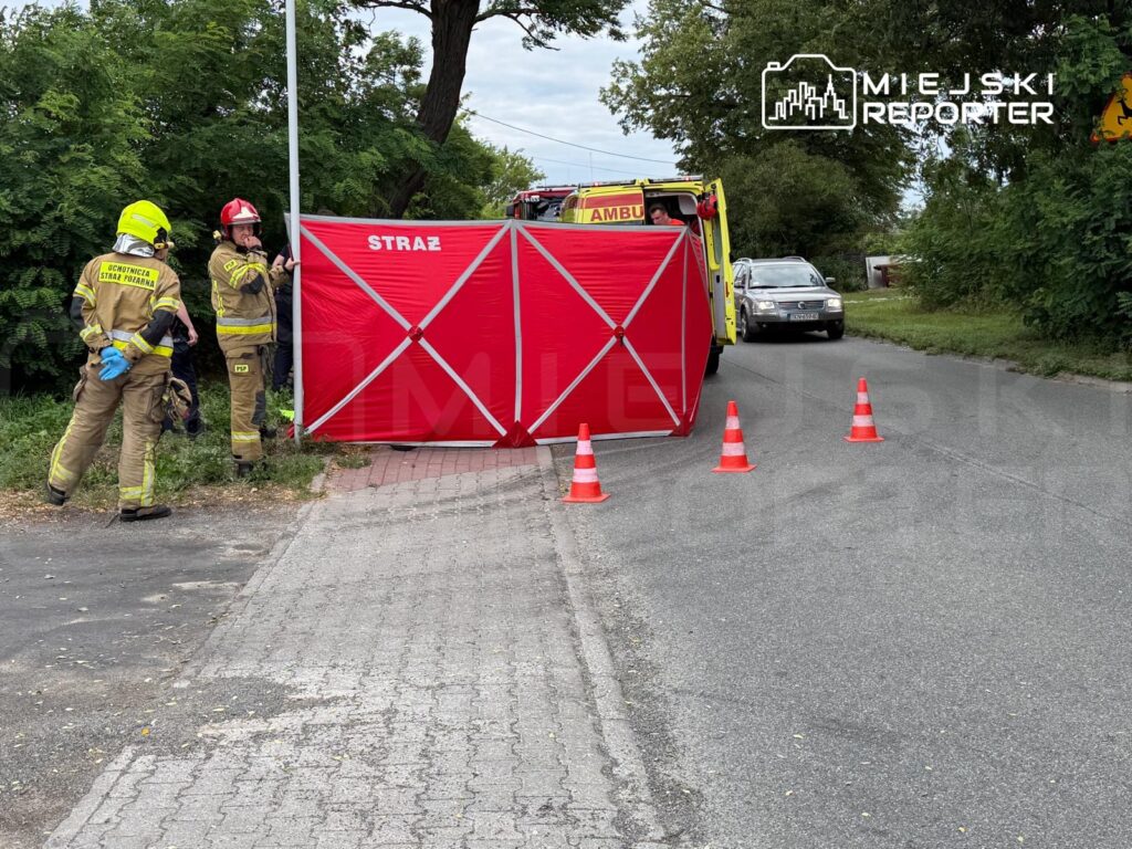 Strażacy w mundurach rozmawiają przy czerwonej bramie z napisem "STRAŻ" na poboczu drogi, obok pojazdu ratunkowego.