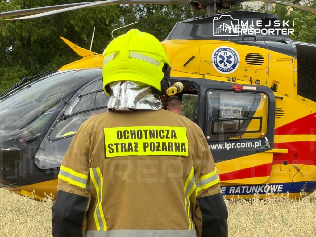 Mężczyzna w żółtej hełmie i mundurze strażackim obserwuje lądowanie śmigłowca ratunkowego na polu.