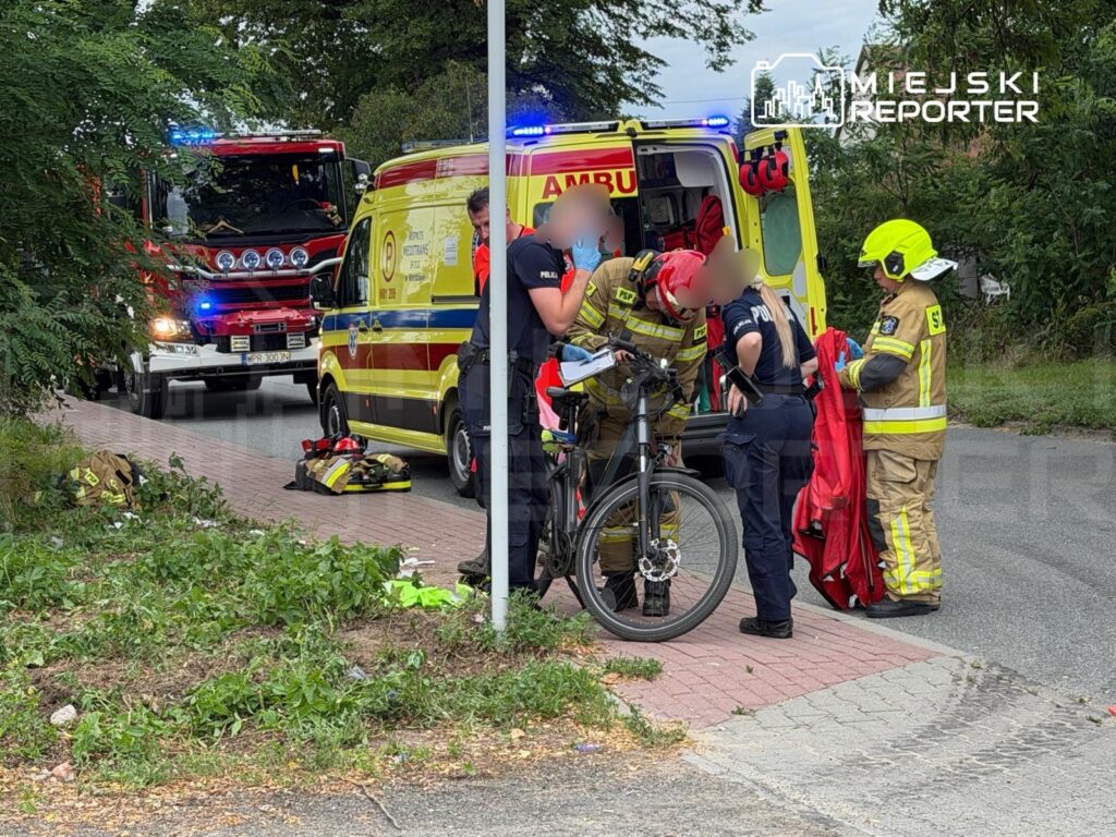 Ratownicy medyczni udzielają pomocy rowerzyście na chodniku, obok stojących pojazdów służb ratunkowych.