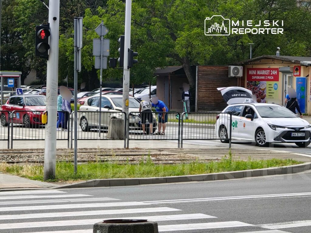 Grupa osób przy samochodzie dostawczym z otwartą klapą bagażnika, w tle zaparkowane auta i sygnalizacja świetlna.