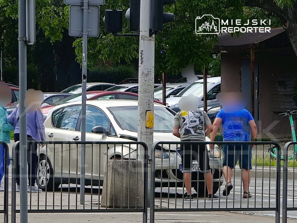 Dwóch mężczyzn zbliża się do białego samochodu na ruchliwej ulicy, obok stojący piesi obserwują sytuację.
