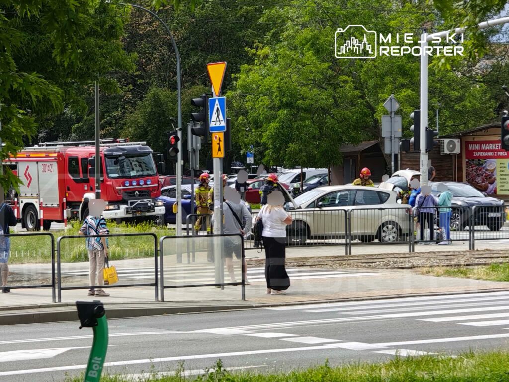 Grupa strażaków w hełmach bada uszkodzony samochód na skrzyżowaniu, obok stojący przechodnie obserwują sytuację.