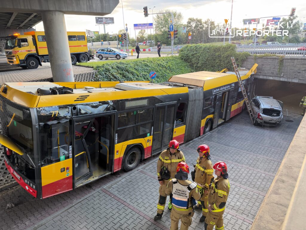 Autobus miejski z uszkodzoną częścią przednią stoi pod wiaduktem, obok zespołu strażaków w hełmach.