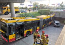 Autobus miejski z uszkodzoną częścią przednią stoi pod wiaduktem, obok zespołu strażaków w hełmach.