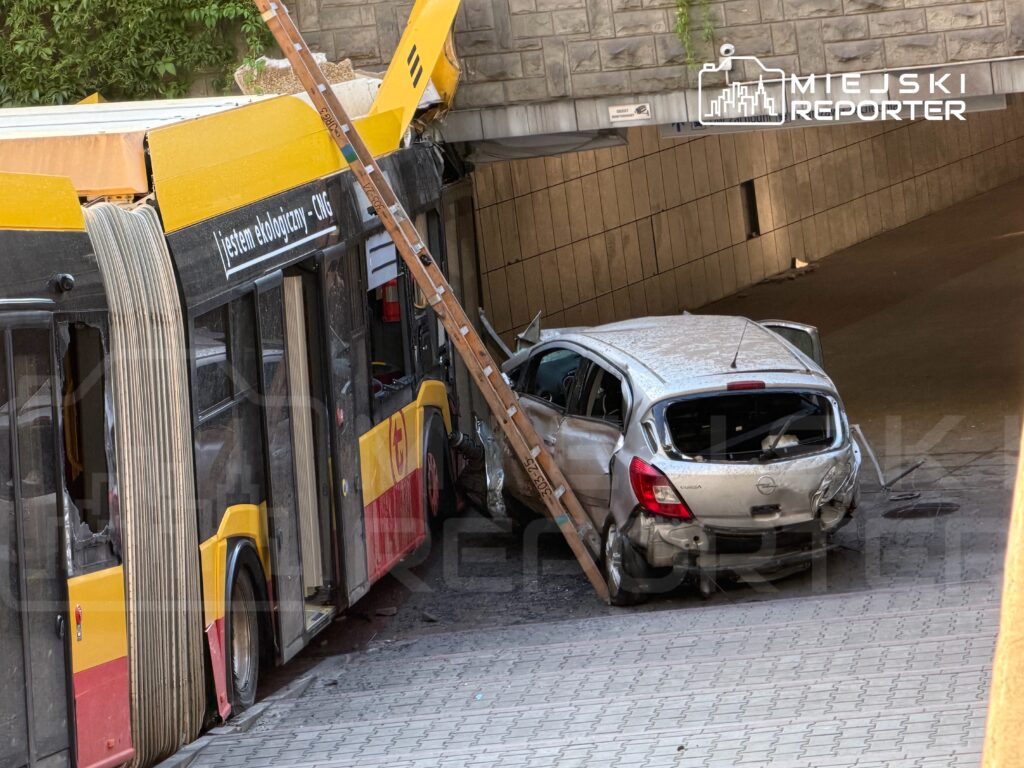 Zniszczony autobus komunikacji miejskiej z uszkodzoną częścią dachu stoi obok rozbitego samochodu osobowego.