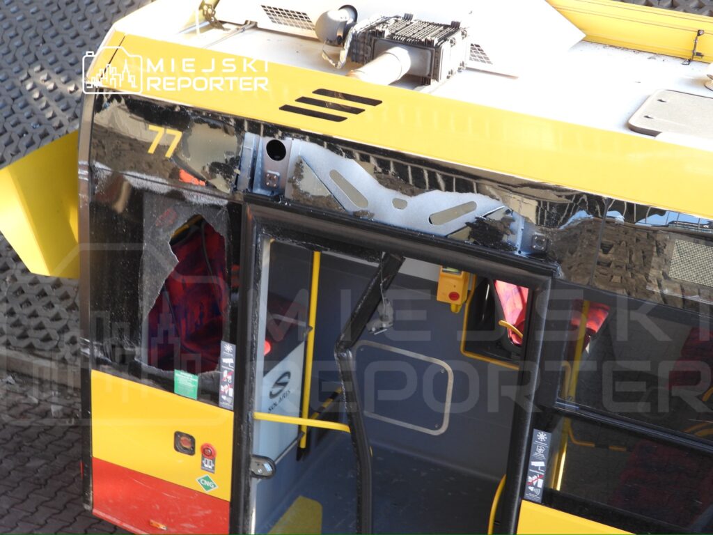 Uszkodzony autobus z wybitym oknem, stojący na płycie dworca, z widocznymi elementami wnętrza.