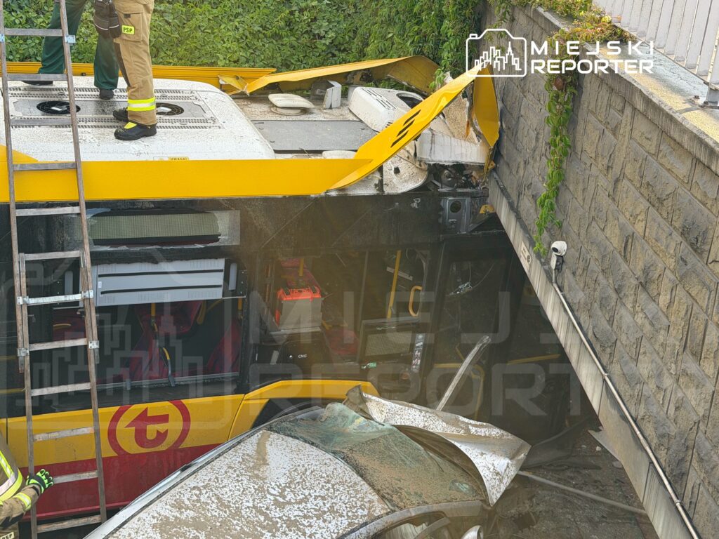 Strażak na dachu autobusu, który uderzył w mur, obok uszkodzony samochód osobowy. W tle widoczna drabina.