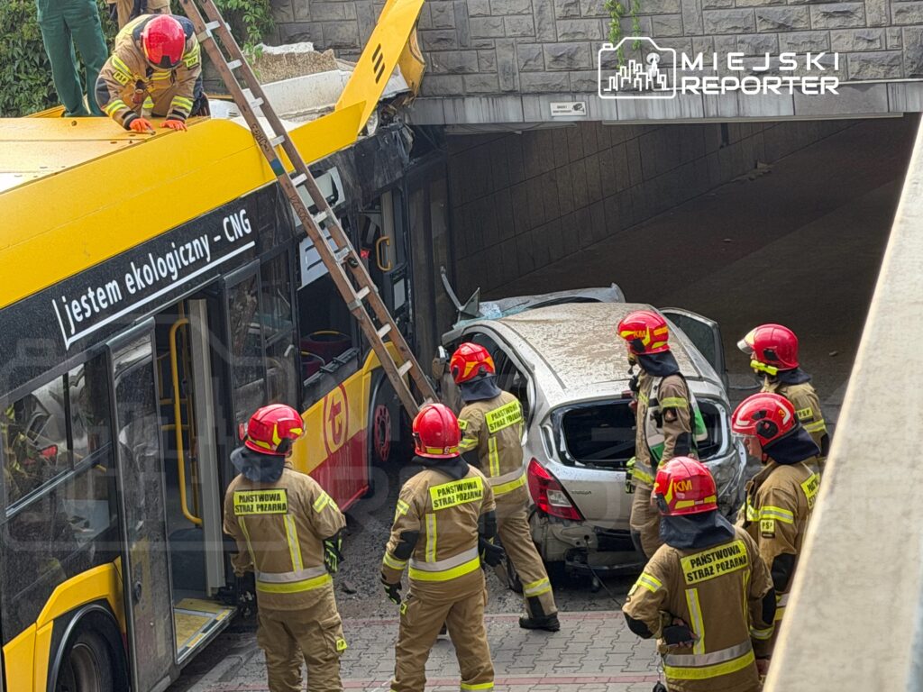 Grupa strażaków w hełmach ocenia uszkodzenia po wypadku, w którym autobus uderzył w osobowy samochód pod wiaduktem.
