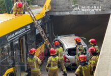 Grupa strażaków w hełmach ocenia uszkodzenia po wypadku, w którym autobus uderzył w osobowy samochód pod wiaduktem.