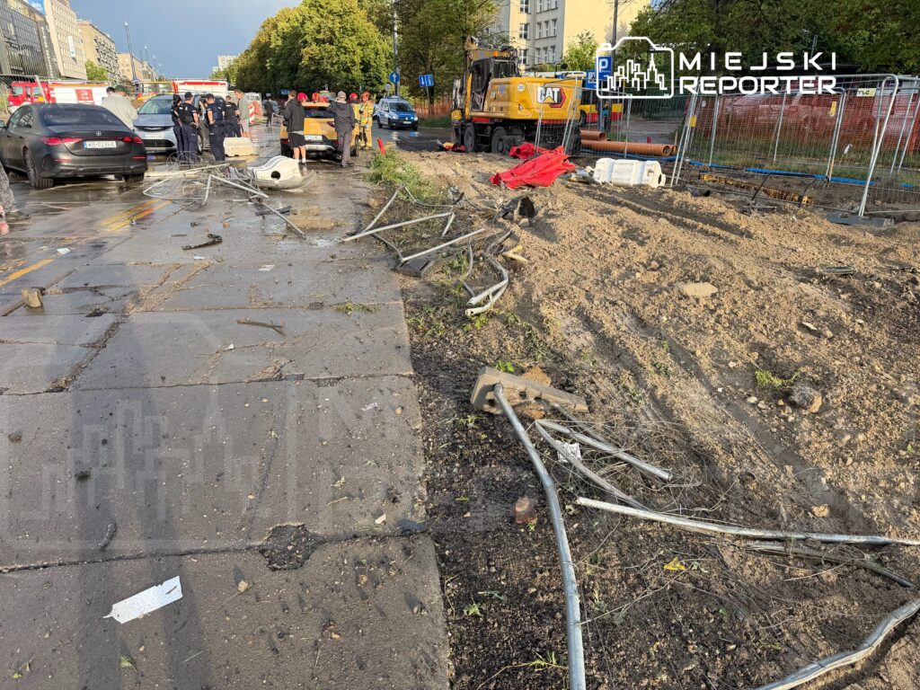Grupa strażaków i policjantów zabezpiecza miejsce wypadku na zniszczonej drodze, obok parkuje samochód ratunkowy.