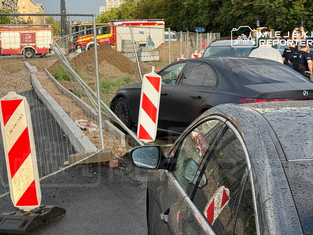 Czarne auto zaparkowane obok oznakowania budowy, w tle widać wozy strażackie i policyjne.