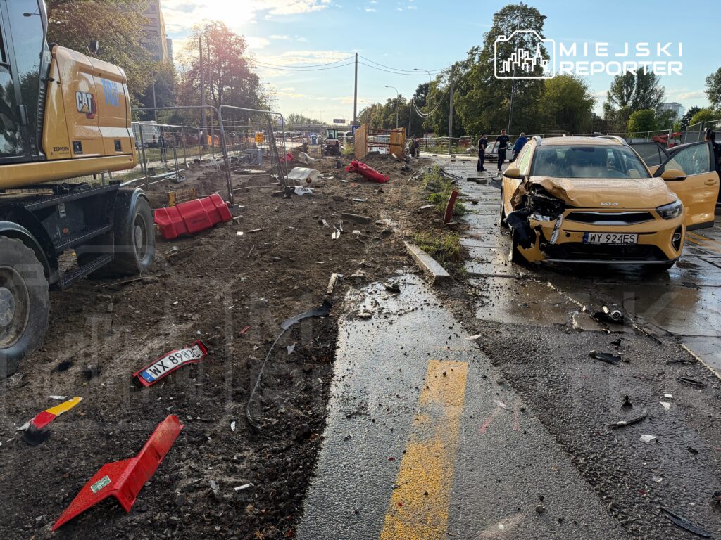 Zniszczony samochód osobowy na drodze, obok sprzęt budowlany i rozkopana nawierzchnia, w tle widoczni pracownicy.