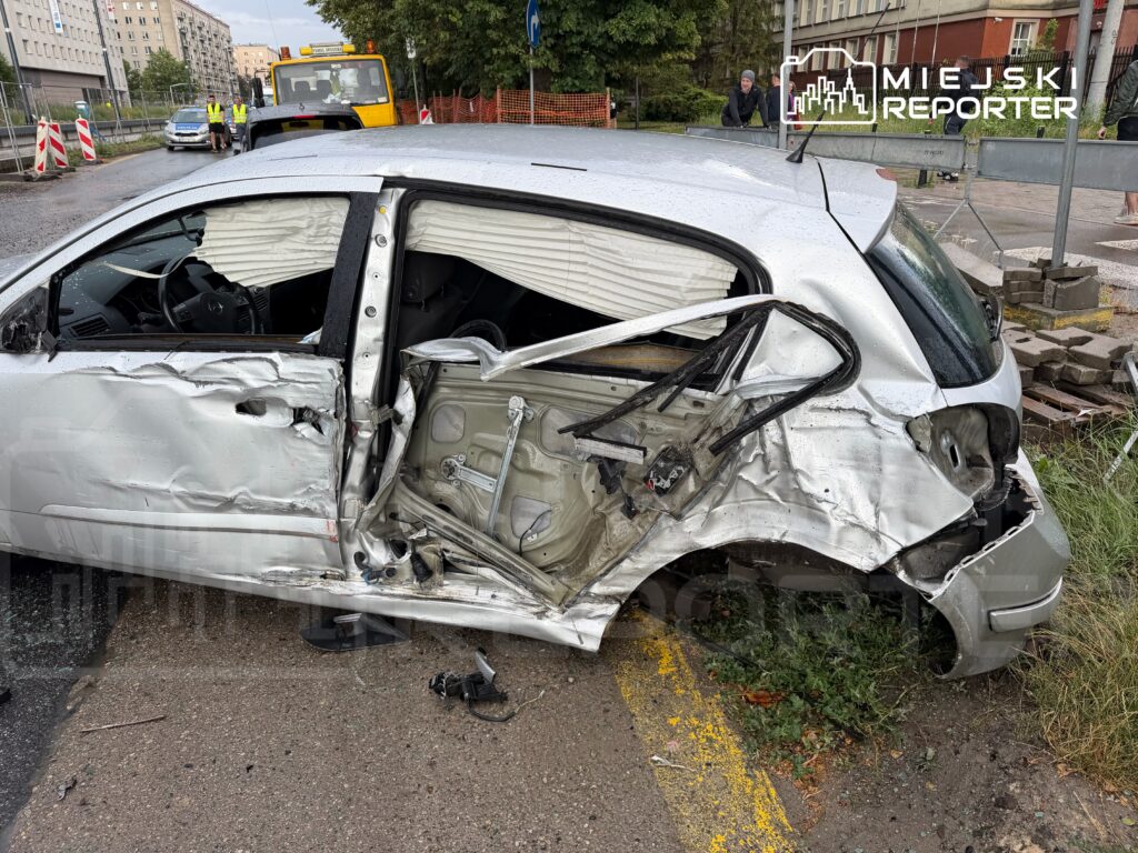 Zniszczony samochód osobowy z wgniecioną stroną stoi na poboczu, obok pracowników w odblaskowych kamizelkach.